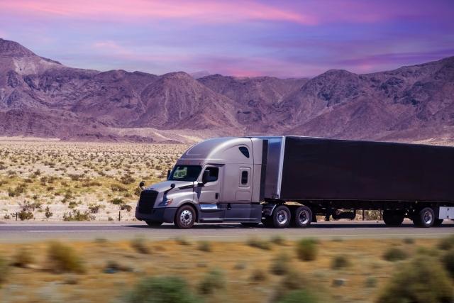 A silver semi-truck with a dark trailer driving on a highway through a desert landscape at dusk, with rugged mountains and a purple-pink sky in the background